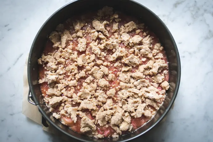 Zubereitung eines veganen Rhabarberkuchens. Streusel werden auf Fruchtfüllung gegeben.
