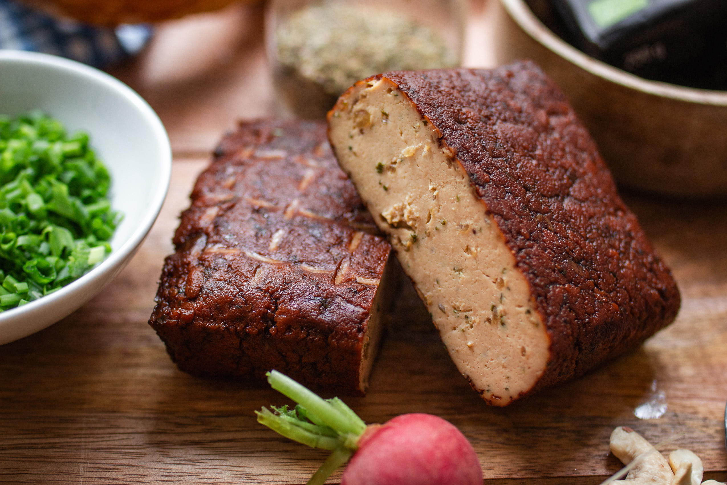 Black Forest Tofu der Marke TAIFUN aufgeschnitten auf einem Holzbrett. Im Anschnitt Radieschen und Schnittlauch.