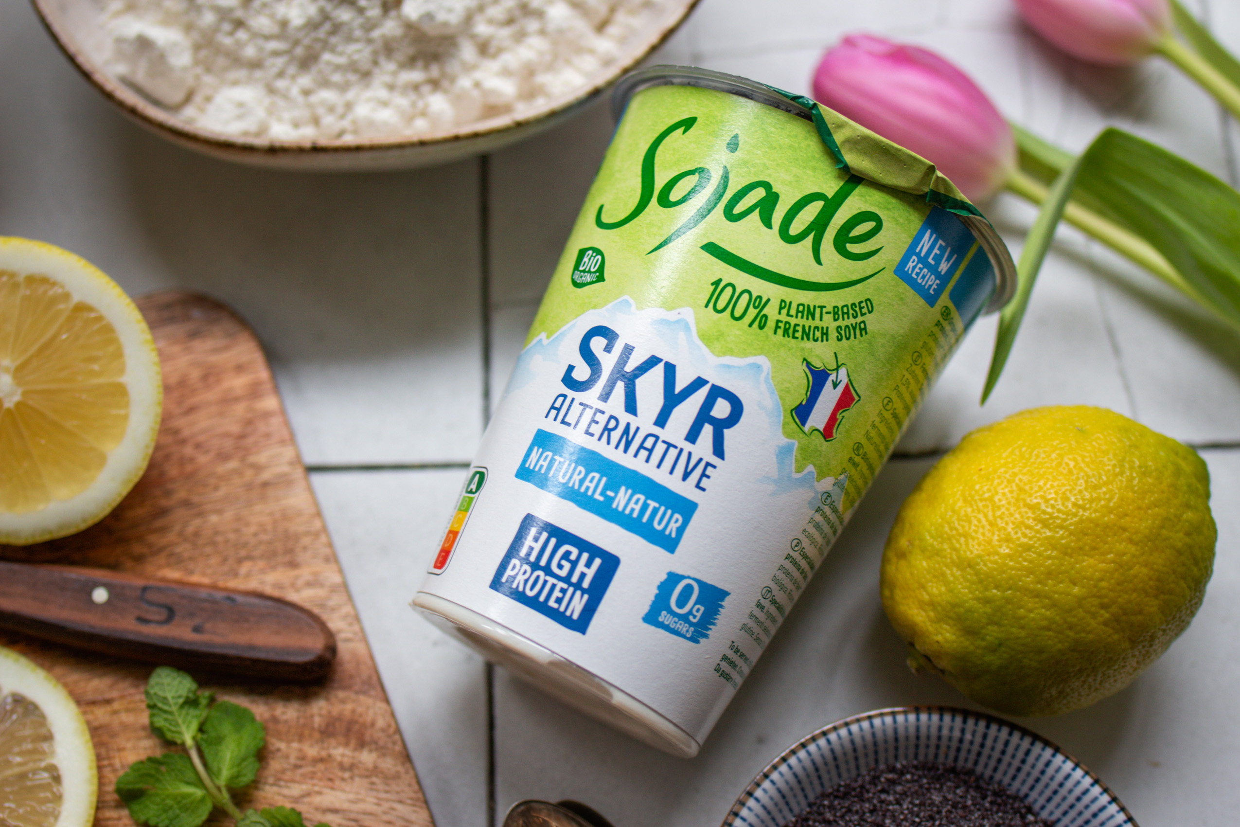 Ein Becher vegane Skyr-Alternative von Sojade auf einem hellen Küchentisch mit Blumen und Zitrone