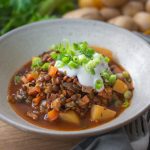 Veganer Berglinseneintopf mit Wurzelgemüse in einer Schale auf einem Tisch mit Kartoffeln und Sellerie im Hintergrund